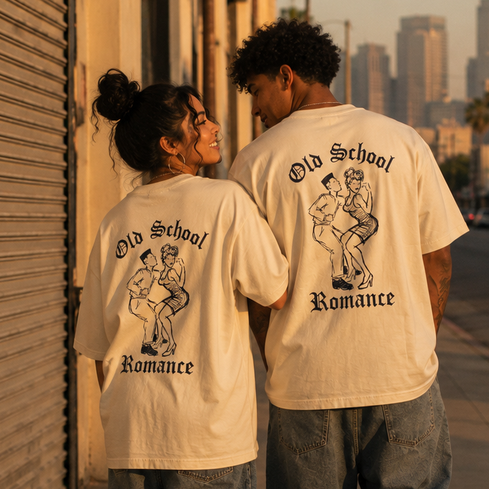 Two people walking away on a city sidewalk at sunset, wearing matching cream graphic T-shirts with a black retro illustration and the words 'Old School Romance' printed on the back, paired with denim shorts.