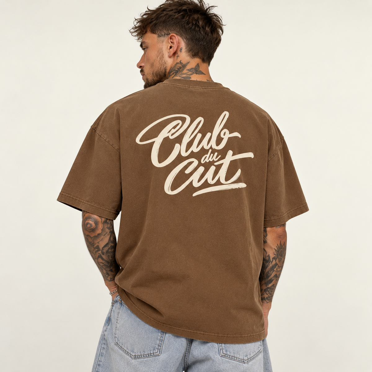 Model seen from behind wearing theCut Club du Cut tee in washed brown, showing the large cream handwritten Club du Cut script across the back.