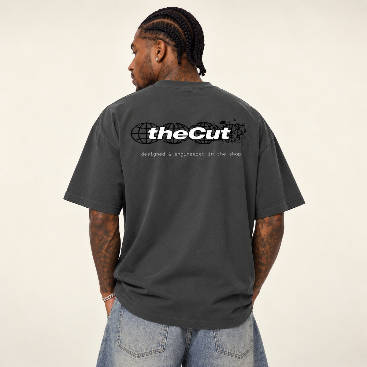 Model seen from behind wearing theCut Globe tee in charcoal grey, showing the large white theCut wordmark and small caption printed across the back.