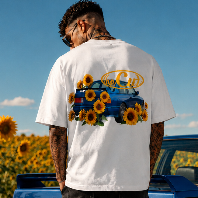 A man stands with his back to the camera in a sunflower field, wearing a white graphic t-shirt with a blue e30 m3 car and sunflower print, black shorts, and white sneakers, with a blue car behind him under a blue sky.