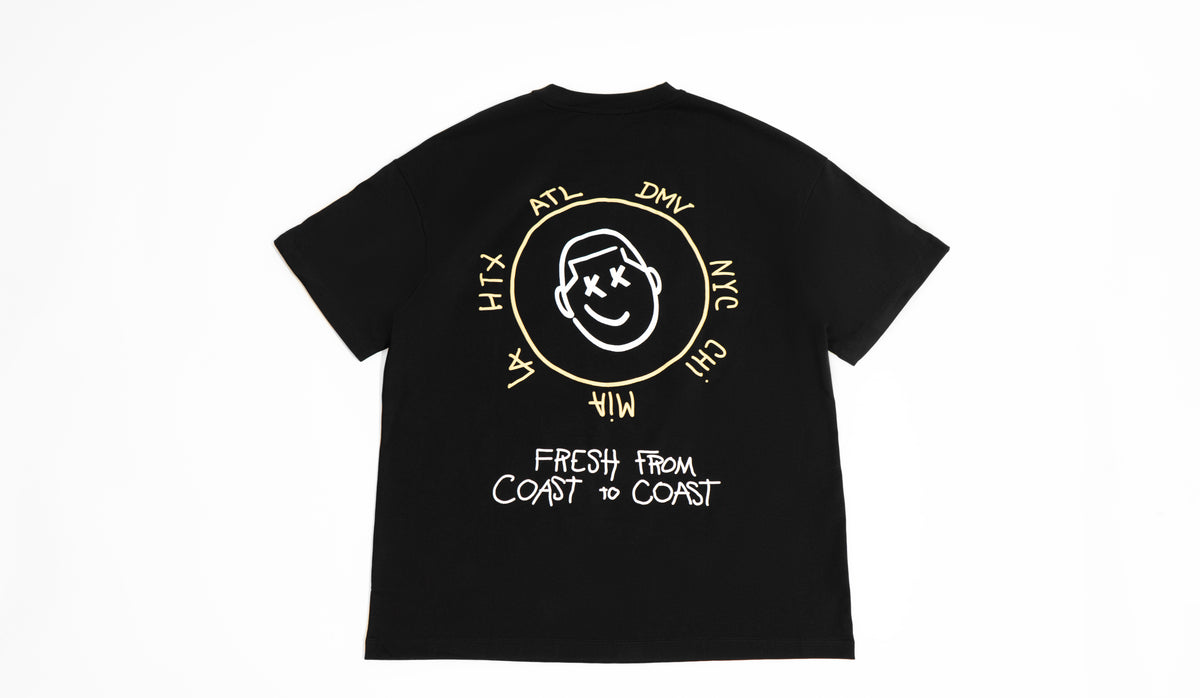 theCut Anywhere tee in black shown flat from the front, with a yellow circular graphic reading Fresh From Coast To Coast.